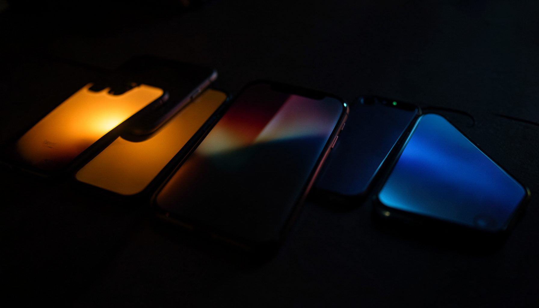 A row of iPhones lying flat on a dark surface, screens glowing softly in warm amber and cool blue tones, moody low-key lighting.