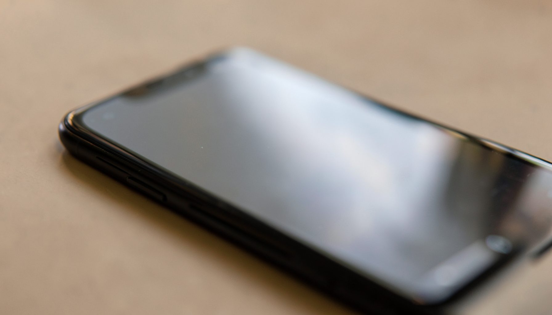 Black smartphone resting on a beige surface with soft warm lighting and a blurred reflective screen.